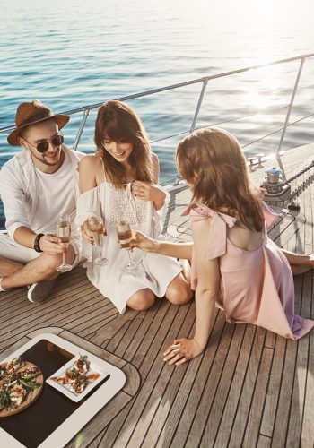 Portrait of three attractive european people sitting on board of yacht and enjoying dinner while drinking champagne and talking cheerfully. Friends worked hard all year to finally enjoy sun and sea. Luxury vacation concept